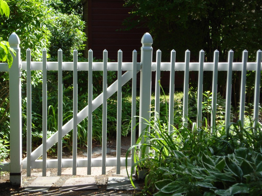 Ein weißer Zaun begrenzt einen Garten, seine Pfosen stehen in Pfostenträgern, die im Boden einbetoniert sind.